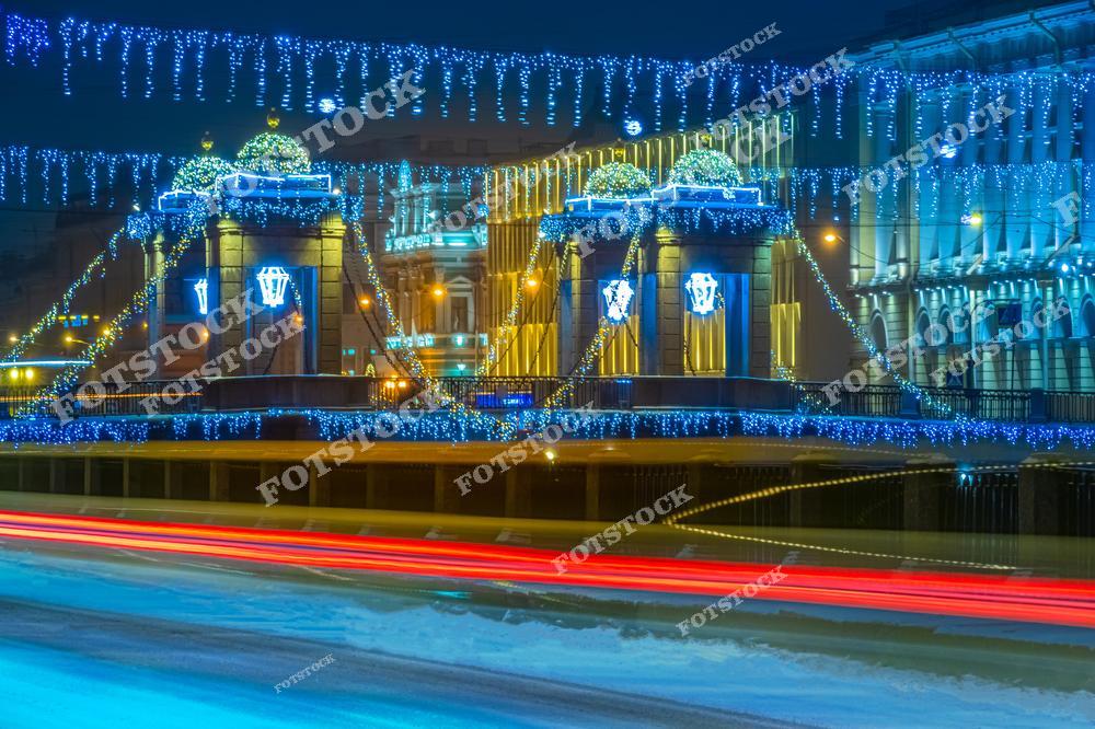 На фотографии изображен мост Ломоносова, украшенный новогодней иллюминацией. Мост расположен в центре Санкт-Петербурга. Яркие гирлянды и световые украшения подчеркивают архитектурные элементы моста и придают ему праздничный вид. Световые следы от проезжающих автомобилей добавляют динамики и подчеркивают ритм жизни города. В зимнюю ночь этот мост через Фонтанку становится ярким символом праздника и волшебства в Петербурге.